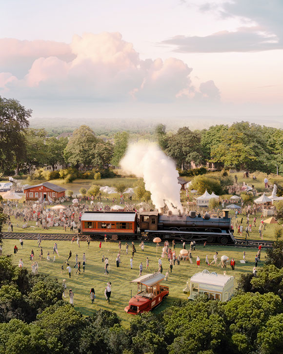 Kép a Normafa tisztásáról egy gőzhajtású vonattal, amelyet vásári hangulatú emberek vesznek körül. A park úgy van kialakítva, hogy befogadó legyen, széles sétányokkal és szabadidős területekkel, amelyek kényelmesen látogathatók minden ember számára.